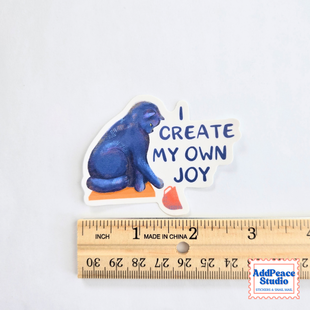 A die-cut sticker featuring a blue cat sitting on an orange block with the text 'I CREATE MY OWN JOY' in blue lettering, shown above a wooden ruler for scale on a white background.