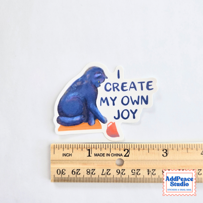 A die-cut sticker featuring a blue cat sitting on an orange block with the text 'I CREATE MY OWN JOY' in blue lettering, shown above a wooden ruler for scale on a white background.