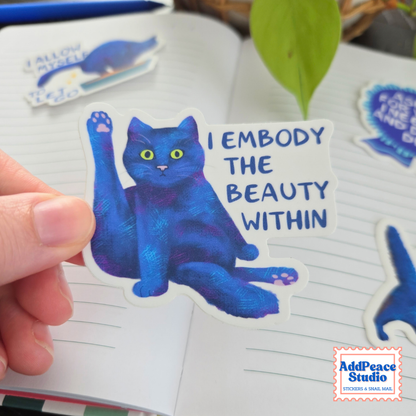 A hand holds a die-cut sticker featuring a blue cat with yellow eyes and the text 'I EMBODY THE BEAUTY WITHIN' next to an open notebook. Other stickers are visible in the background, and a 'AddPeace Studio' logo is in the bottom right corner.