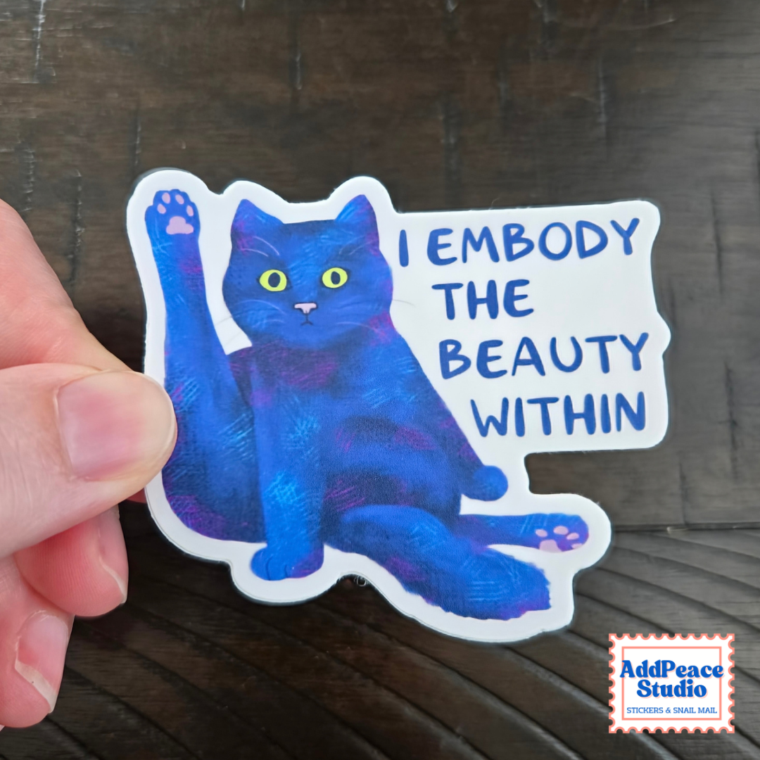 A hand holds a die-cut sticker featuring a blue cat with yellow eyes and the text 'I EMBODY THE BEAUTY WITHIN' on a dark wood background. An AddPeace Studio logo is visible in the bottom right corner.