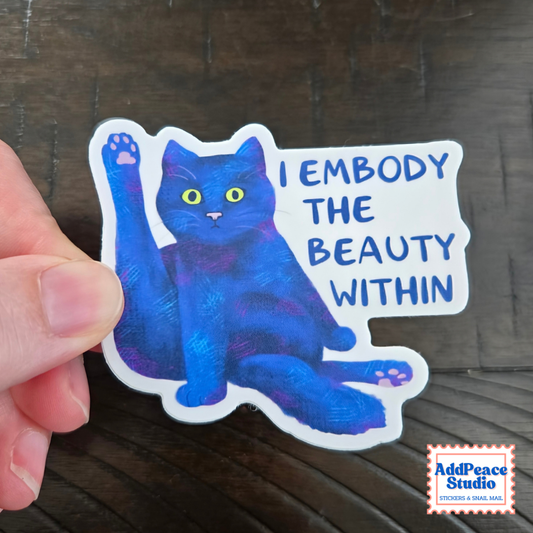 A hand holds a die-cut sticker featuring a blue cat with yellow eyes and the text 'I EMBODY THE BEAUTY WITHIN' on a dark wood background. An AddPeace Studio logo is visible in the bottom right corner.