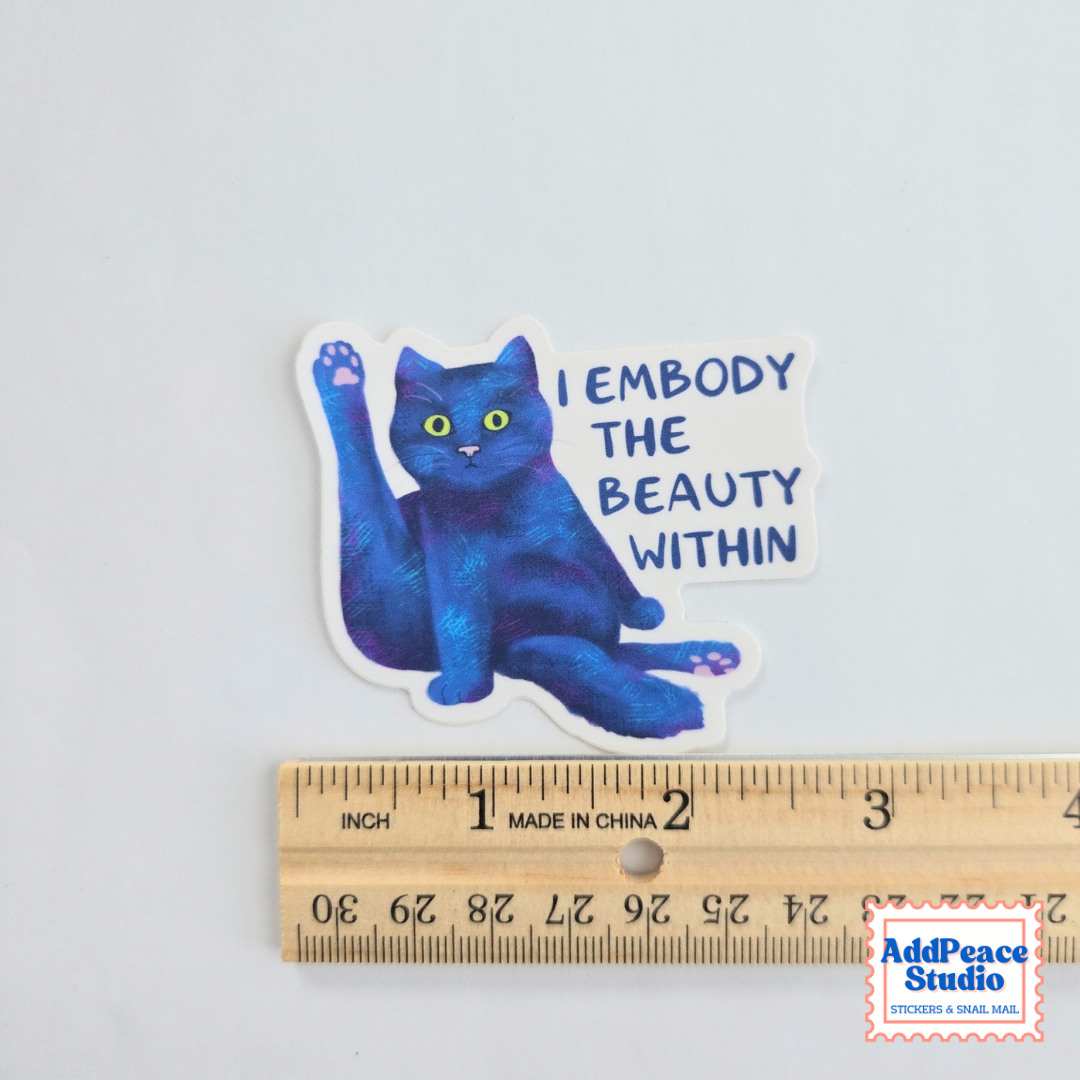 A die-cut sticker featuring a blue cat with a raised paw and the text 'I EMBODY THE BEAUTY WITHIN' in blue lettering, shown above a wooden ruler for scale on a white background.