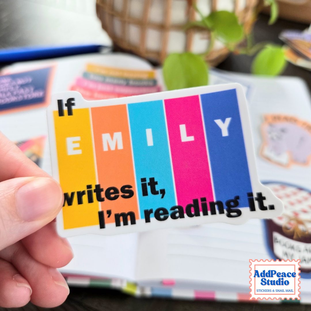 A hand holds a die-cut sticker shaped like a row of books featuring the text 'If EMILY writes it, I'm reading it.' in black over vertical color blocks of yellow, orange, teal, pink, and blue.