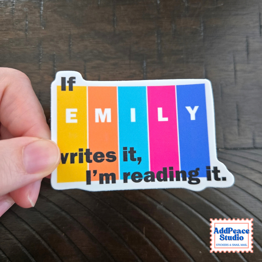 A hand holds a die-cut sticker shaped like a row of books featuring vertical color blocks in yellow, orange, blue, pink, and purple with the text 'If EMILY writes it, I'm reading it.' on a dark wood background.