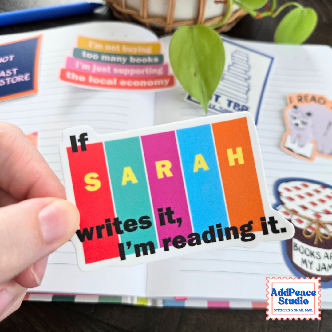 A hand holds a sticker that says If Sarah writes it I'm reading it. The background is an open notebook with stickers and plants.
