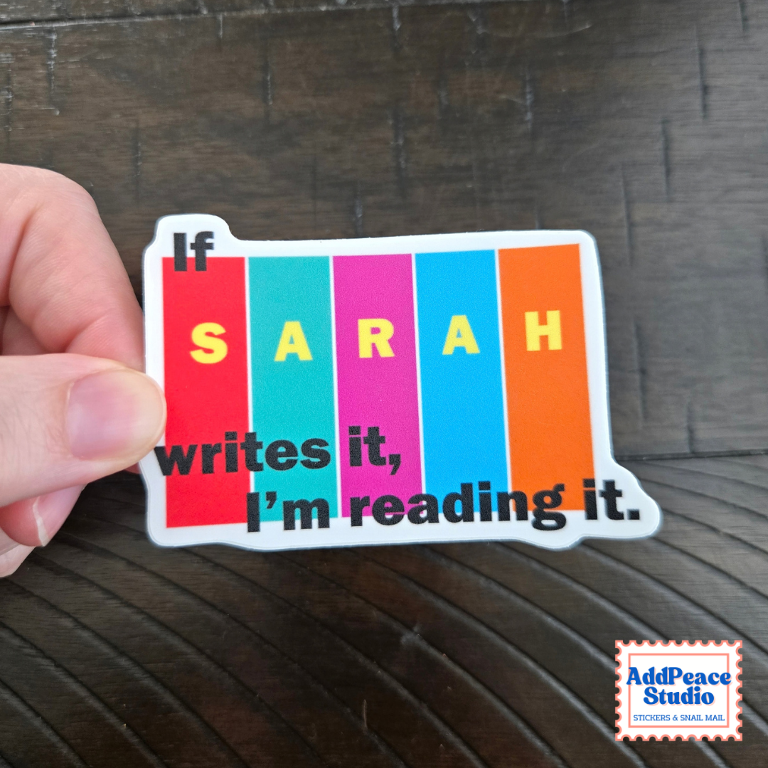 A hand holds a die-cut sticker that reads 'If SARAH writes it, I’m reading it.' over vertical rainbow-colored books.