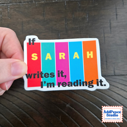 A hand holds a die-cut sticker that reads 'If SARAH writes it, I’m reading it.' over vertical rainbow-colored books.