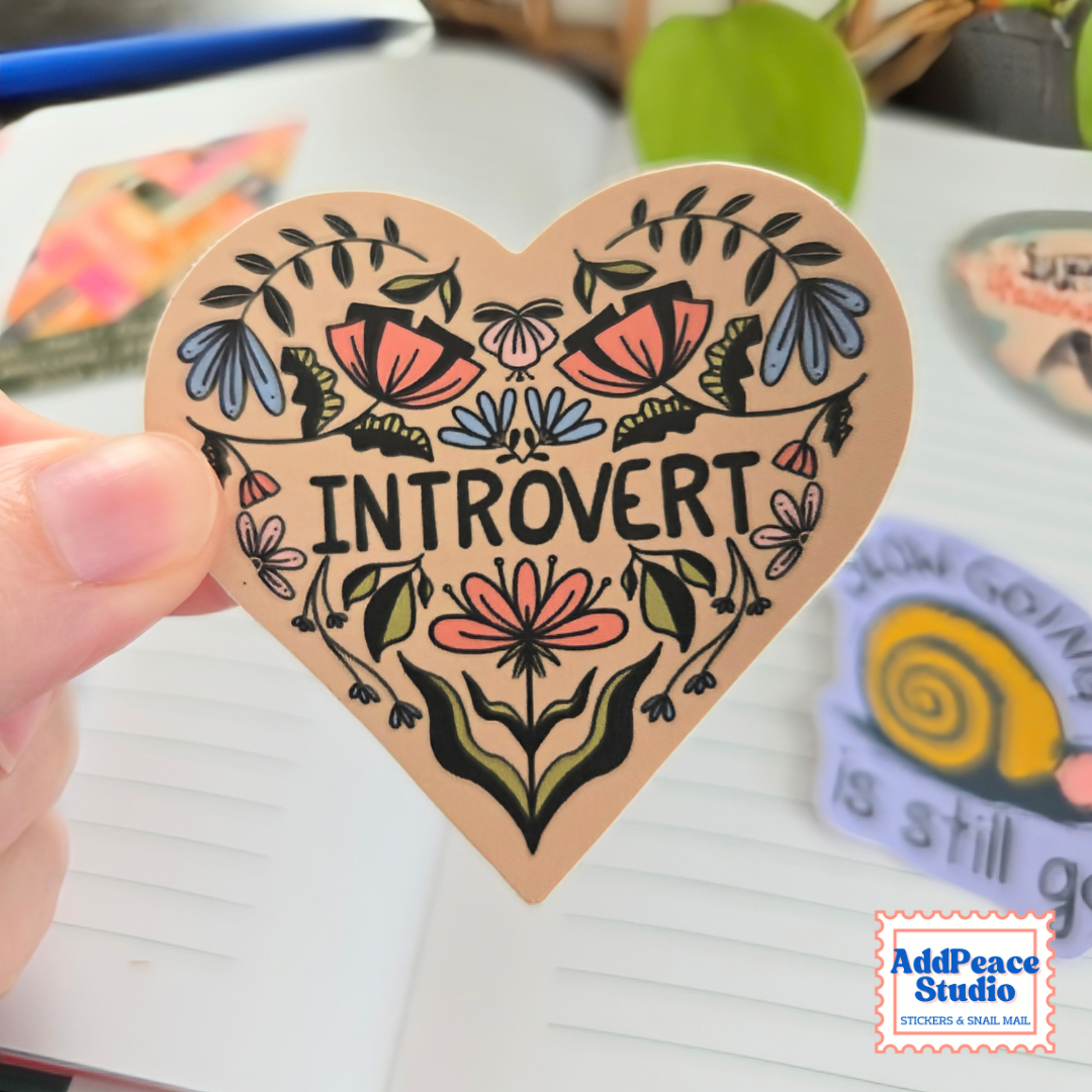 A hand holds a heart-shaped sticker featuring the word 'INTROVERT' surrounded by colorful floral illustrations. The sticker has a warm beige background with black text and black outlines, with pink, blue, and green accents in the artwork.