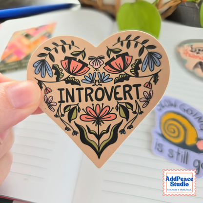 A hand holds a heart-shaped sticker featuring the word 'INTROVERT' surrounded by colorful floral illustrations. The sticker has a warm beige background with black text and black outlines, with pink, blue, and green accents in the artwork.