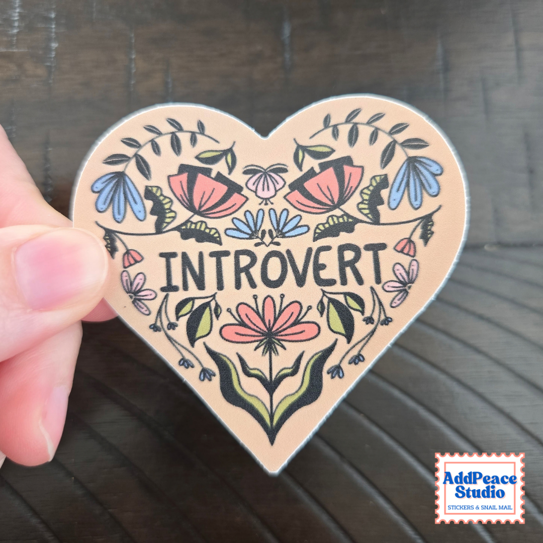 A hand holds a heart-shaped sticker with the word 'INTROVERT' surrounded by colorful floral illustrations on a dark wood background. The AddPeace Studio logo is visible in the bottom right corner.