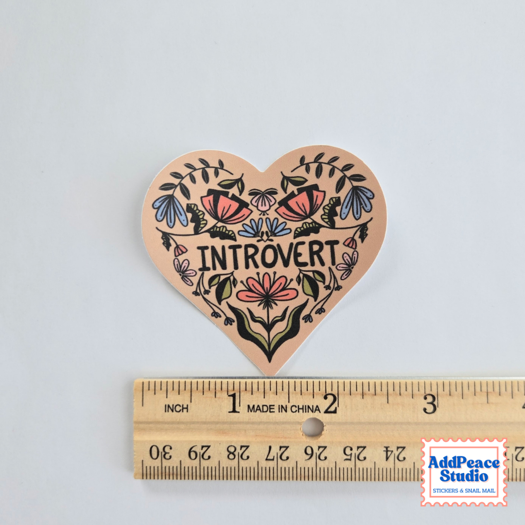 A heart-shaped sticker with a colorful floral design and the word 'INTROVERT' in the center, shown above a wooden ruler for scale on a light gray background.