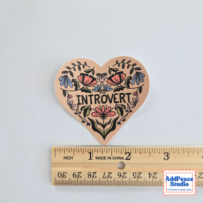 A heart-shaped sticker with a colorful floral design and the word 'INTROVERT' in the center, shown above a wooden ruler for scale on a light gray background.