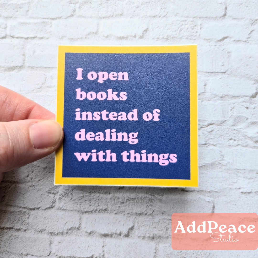 Hand holding a sticker with text 'I open books instead of dealing with things' against a textured white background.