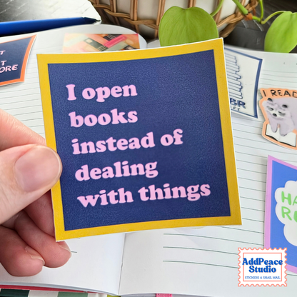 A hand holds a square sticker with a navy blue background and yellow border, featuring the text 'I open books instead of dealing with things' in pink. The sticker is shown over an open notebook with other stickers and books in the background.