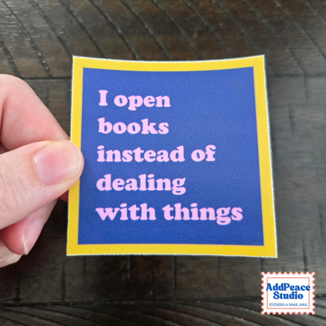 A hand holds a square sticker with a blue background, yellow border, and pink text that reads, 'I open books instead of dealing with things.' The sticker is photographed over a dark wood surface, with an AddPeace Studio logo in the bottom right corner.
