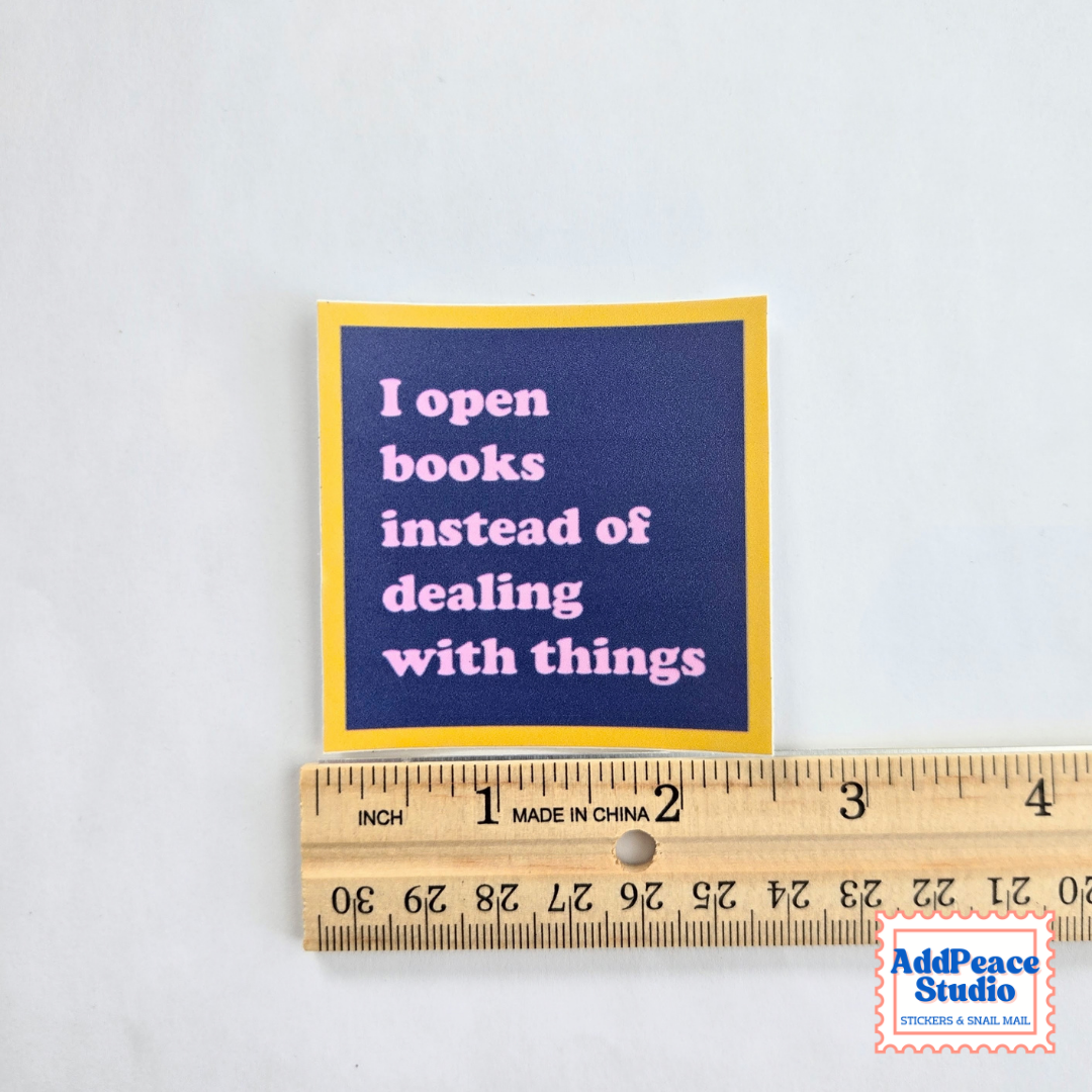 A square sticker with a navy blue background and yellow border, featuring the quote 'I open books instead of dealing with things' in pink text. The sticker is shown next to a wooden ruler to indicate its size, on a light gray background.