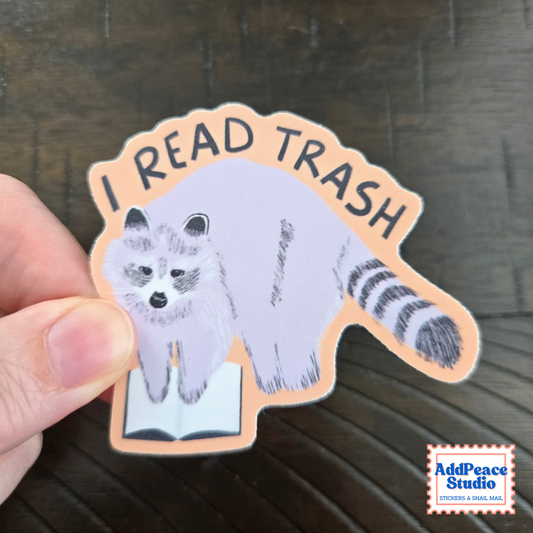 A hand holds a die-cut sticker featuring a gray and white raccoon reading an open book, with the text 'I READ TRASH' arched above. The sticker has a peach border and is photographed over a dark wood surface.