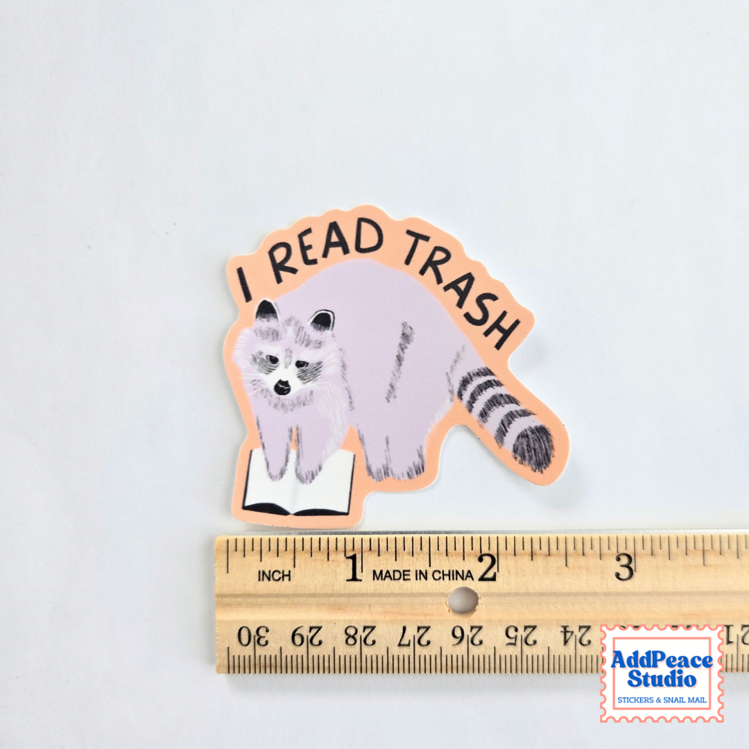 A die-cut sticker featuring a raccoon reading a book with the text 'I READ TRASH' in black lettering, shown above a wooden ruler for scale on a white background.