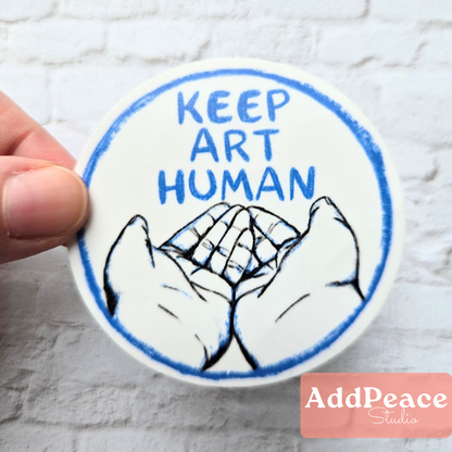 Circular sticker with 'Keep Art Human' text and hands holding a heart, held by a hand on a textured white background.
