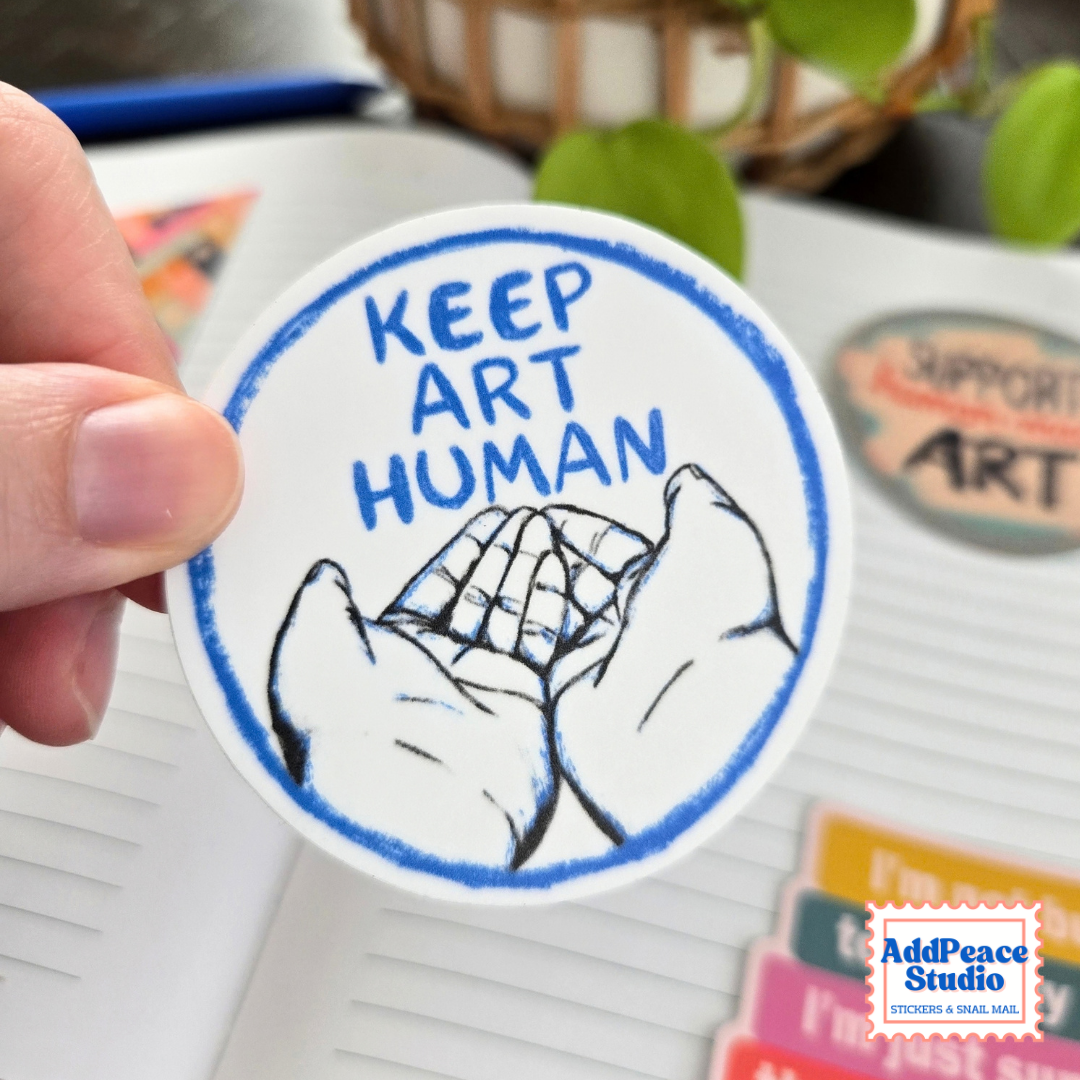 A hand holds a round white sticker with blue and black artwork and the text 'KEEP ART HUMAN.' The background shows a notebook, colorful stationery, and a blurred plant.