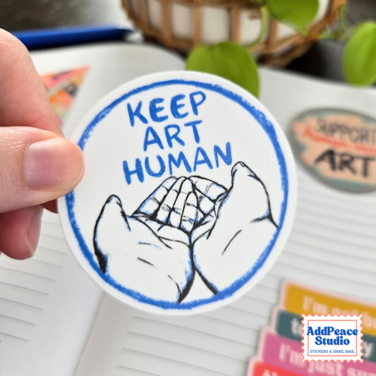 A hand holds a round white sticker with blue and black artwork and the text 'KEEP ART HUMAN.' The background shows a notebook, colorful stationery, and a blurred plant.