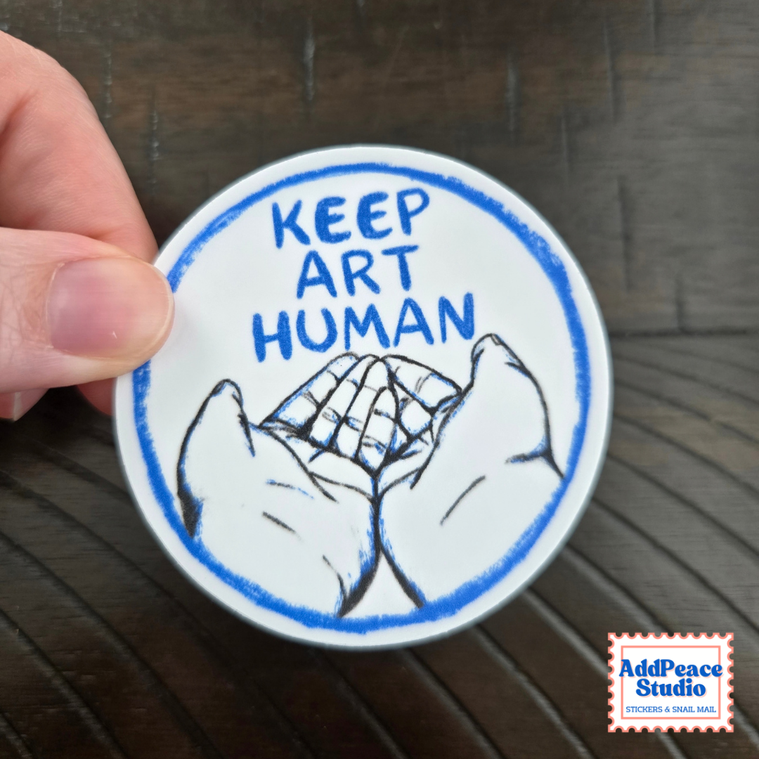 A round white sticker held by a hand over a dark wood background. The sticker features the text 'KEEP ART HUMAN' in blue lettering inside a blue circle, with a black and white illustration of open hands.