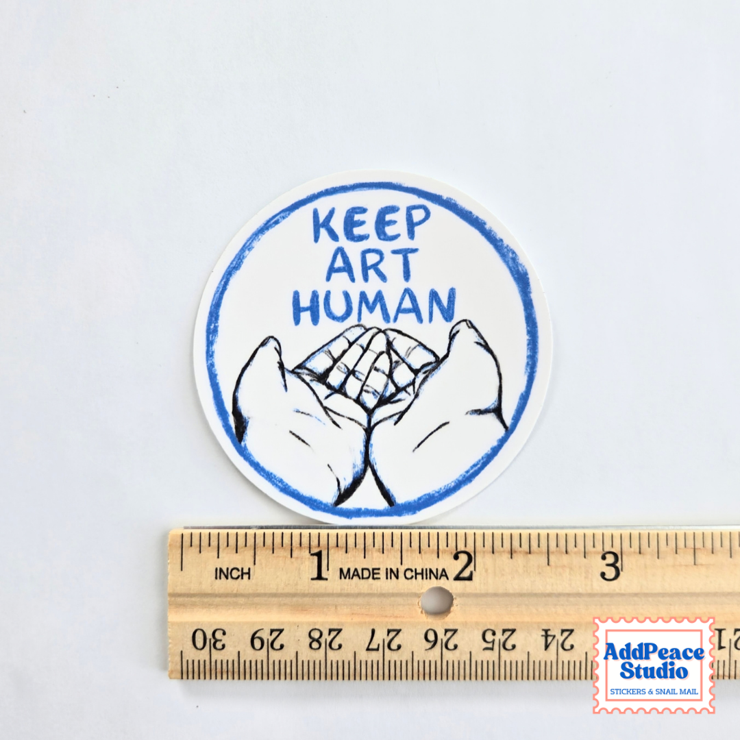 A round sticker with the text 'KEEP ART HUMAN' and a drawing of two open hands, shown above a wooden ruler for scale on a white background.