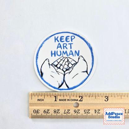 A round sticker with the text 'KEEP ART HUMAN' and a drawing of two open hands, shown above a wooden ruler for scale on a white background.