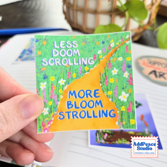A hand holds a square sticker featuring a colorful illustration of a winding orange path through a green field of flowers, with the text 'LESS DOOM SCROLLING' and 'MORE BLOOM STROLLING'.