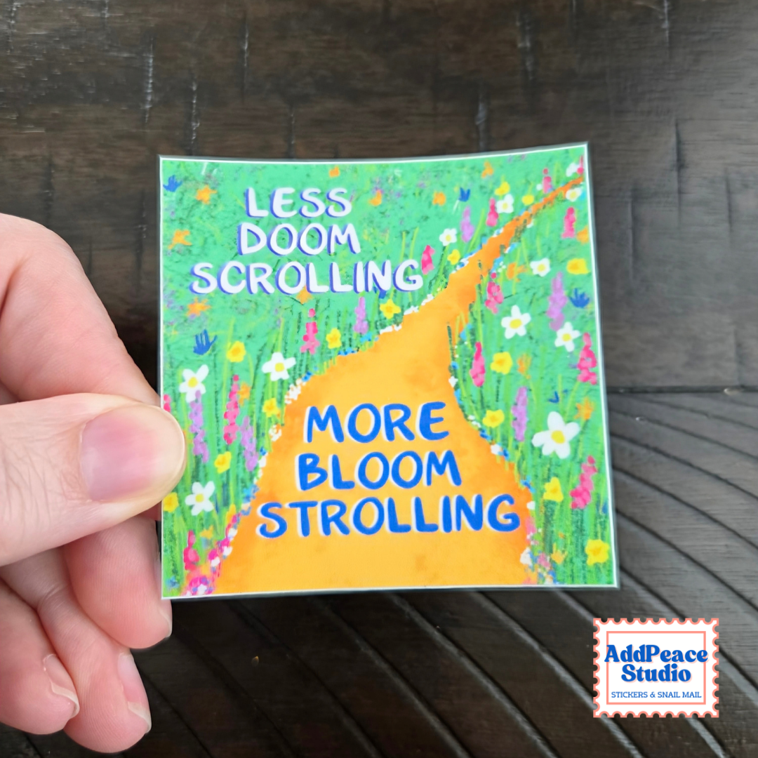 A hand holds a square sticker featuring a colorful illustration of a winding orange path through a green field of flowers, with the text 'LESS DOOM SCROLLING' at the top and 'MORE BLOOM STROLLING' on the path. The background is a dark wooden surface, and an 'AddPeace Studio' logo watermark is in the bottom right corner.