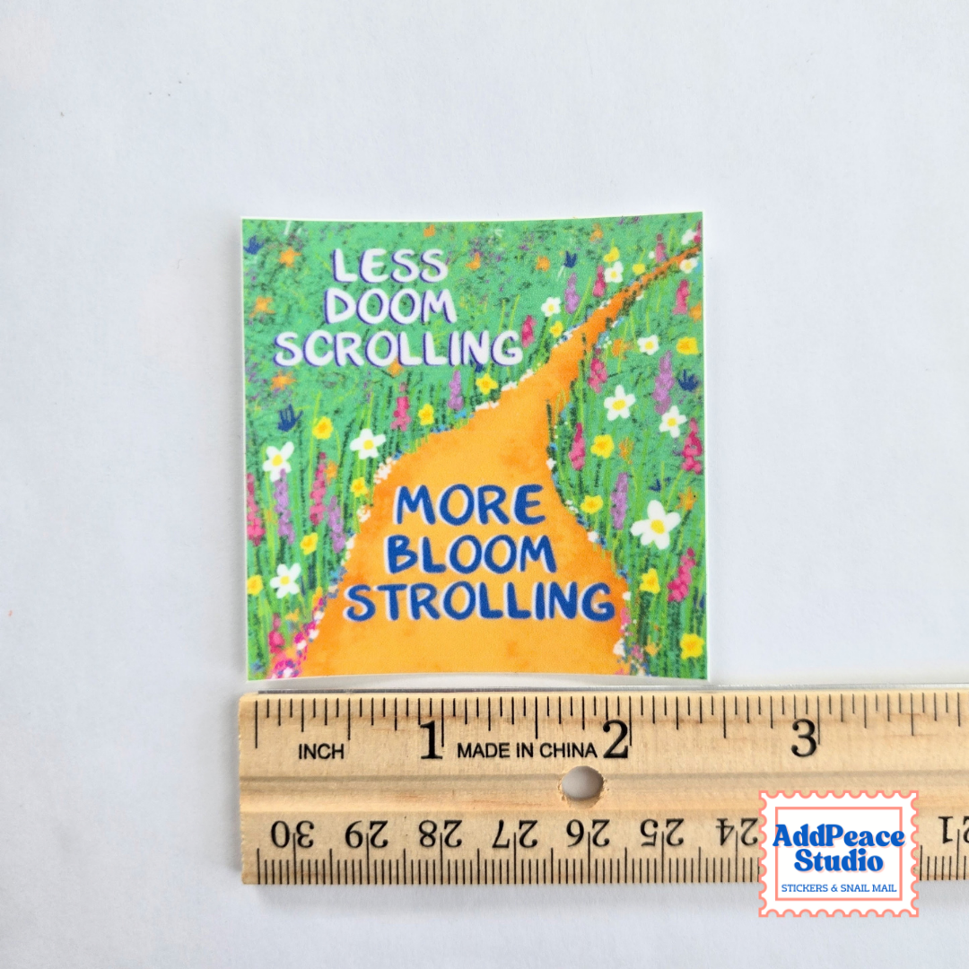 A square printed card or sticker with a colorful floral path illustration and the text 'LESS DOOM SCROLLING MORE BLOOM STROLLING' is shown above a wooden ruler for scale on a white background. An AddPeace Studio watermark appears in the bottom right corner.