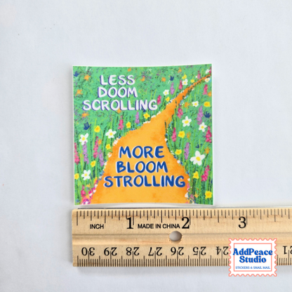 A square printed card or sticker with a colorful floral path illustration and the text 'LESS DOOM SCROLLING MORE BLOOM STROLLING' is shown above a wooden ruler for scale on a white background. An AddPeace Studio watermark appears in the bottom right corner.