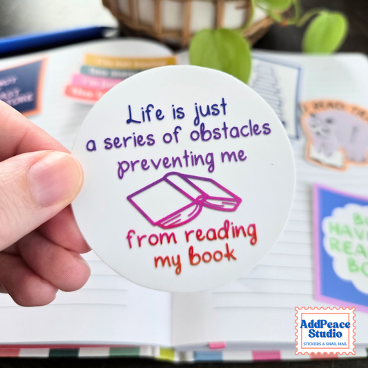 A hand holds a round white sticker with a purple and red quote that reads 'Life is just a series of obstacles preventing me from reading my book' and a simple book illustration. The background shows an open notebook with colorful sticky notes and a blurred plant.