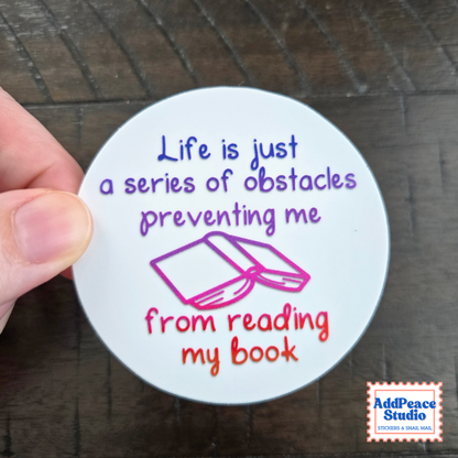 A hand holds a round white sticker with the quote 'Life is just a series of obstacles preventing me from reading my book' in purple and orange lettering, with a pink book illustration. The background is a dark wood surface, and an 'AddPeace Studio' logo is in the bottom right corner.