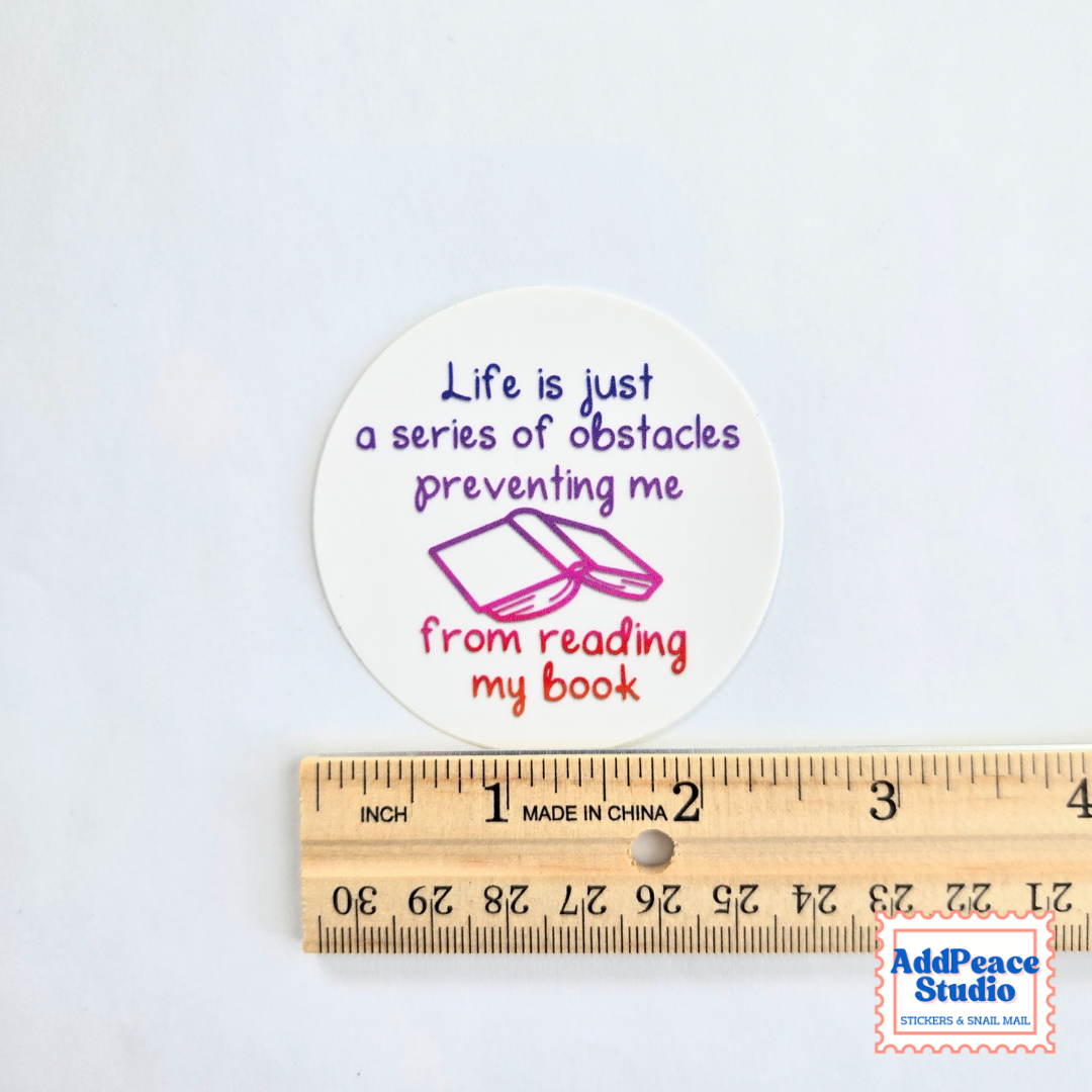 A round white sticker with a reading-themed quote in purple and red text and a small book illustration, shown above a wooden ruler for scale. The sticker reads: 'Life is just a series of obstacles preventing me from reading my book.