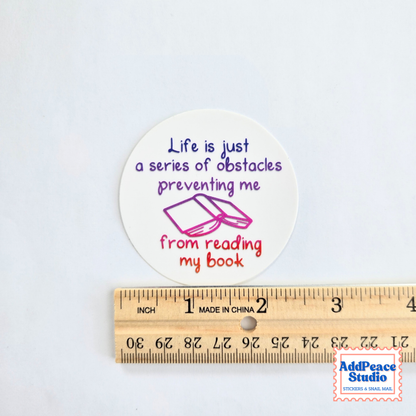 A round white sticker with a reading-themed quote in purple and red text and a small book illustration, shown above a wooden ruler for scale. The sticker reads: 'Life is just a series of obstacles preventing me from reading my book.