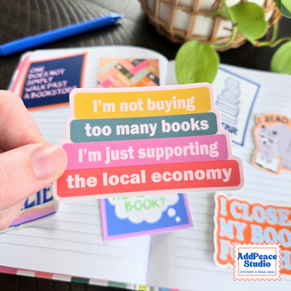 A hand holds a stack of colorful sticker labels with book-themed text, with an open notebook and other stickers in the background. The AddPeace Studio logo is visible in the bottom right corner.