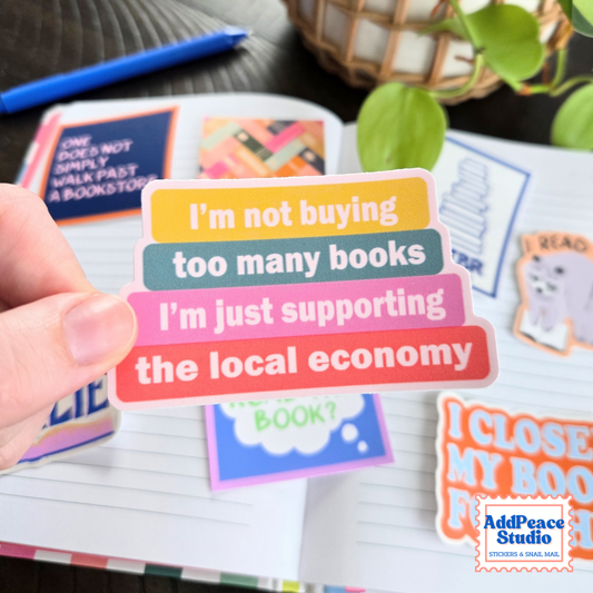 A hand holds a stack of colorful sticker labels with book-themed text, with an open notebook and other stickers in the background. The AddPeace Studio logo is visible in the bottom right corner.