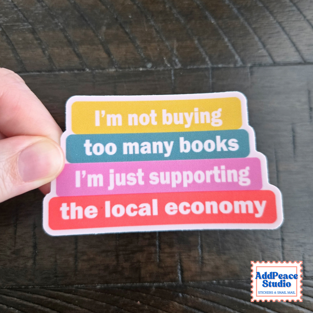 A hand holds a colorful sticker with stacked text reading, 'I'm not buying too many books I'm just supporting the local economy.' The sticker has yellow, teal, pink, and coral sections with white text.