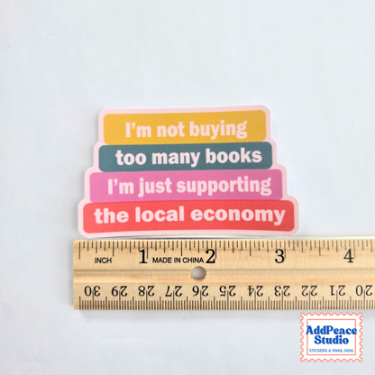 A small multicolor sticker with stacked book shapes and the text “I’m not buying too many books I’m just supporting the local economy” is shown above a wooden ruler to indicate size. The ruler shows the sticker is approximately 2 inches tall.