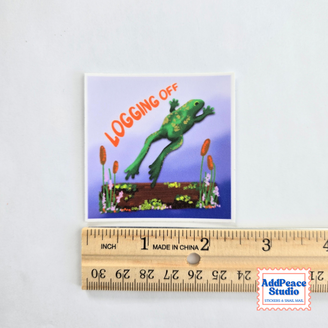 A square sticker featuring a green frog jumping over a garden bed with the text 'LOGGING OFF' in orange, displayed above a wooden ruler for scale on a white background.