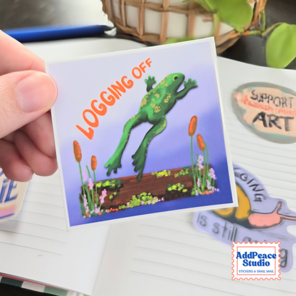 A hand holds a square sticker featuring a green frog jumping over a log with the text 'LOGGING OFF' on a light blue background. The sticker is shown in a desk setting with other stickers and a notebook visible.