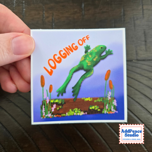 A hand holds a square sticker featuring a green frog jumping over a log with the words 'LOGGING OFF' in orange, set against a blue background with small plants. The AddPeace Studio logo appears in the bottom right corner.