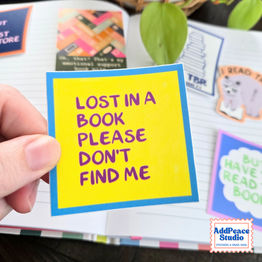A hand holds a yellow and blue sticker that reads 'LOST IN A BOOK PLEASE DON'T FIND ME' in purple lettering, set against an open notebook with other colorful book-themed stickers.