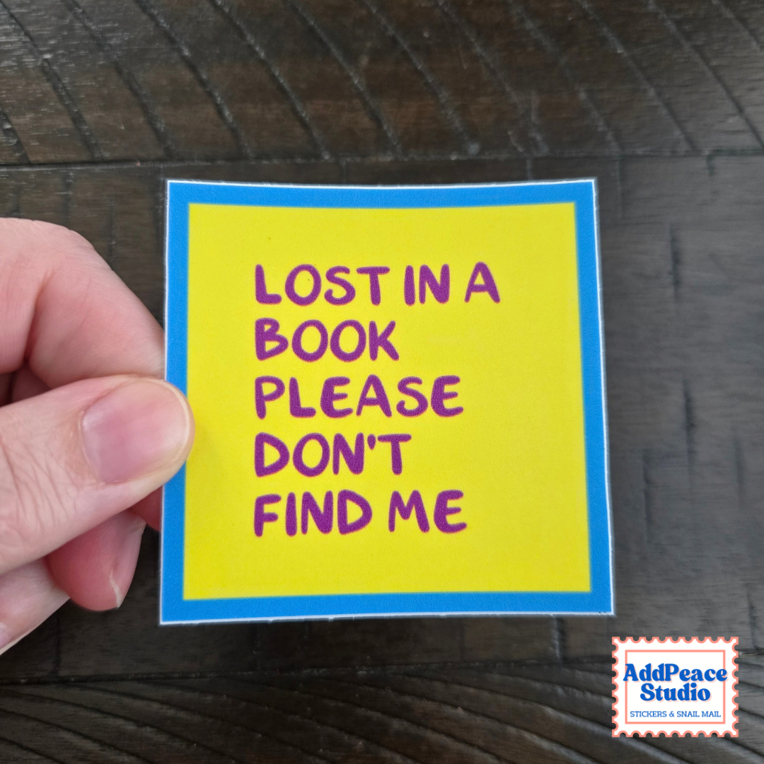 A hand holds a square yellow sticker with a blue border and purple text that reads 'LOST IN A BOOK PLEASE DON'T FIND ME' against a dark wood background. An AddPeace Studio logo appears in the bottom right corner.