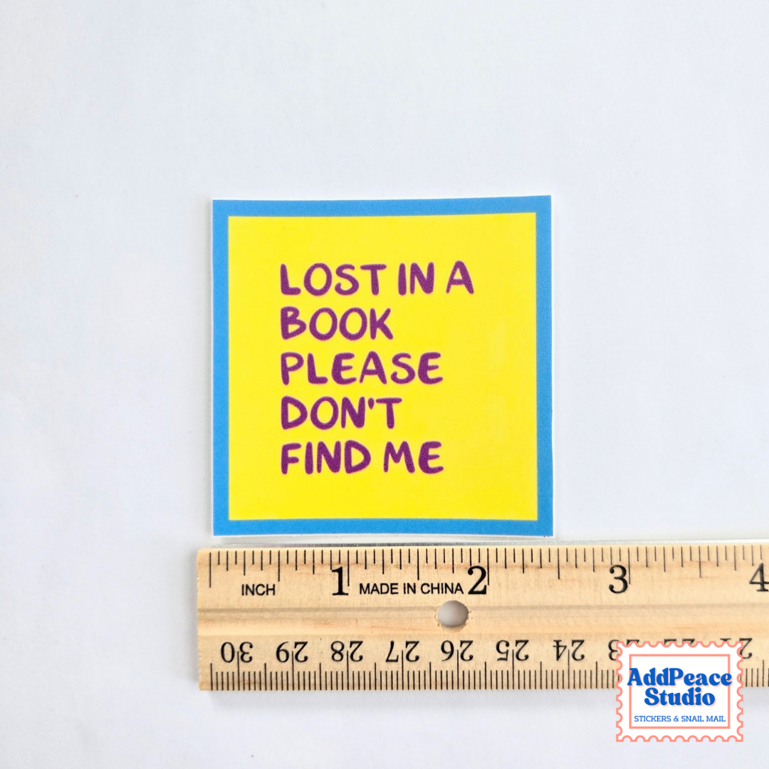 A square yellow sticker with a blue border and purple text reading 'LOST IN A BOOK PLEASE DON'T FIND ME' is shown above a wooden ruler on a white background. An AddPeaceStudio watermark appears in the bottom right corner.