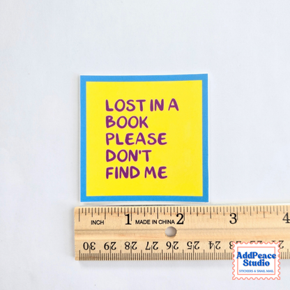 A square yellow sticker with a blue border and purple text reading 'LOST IN A BOOK PLEASE DON'T FIND ME' is shown above a wooden ruler on a white background. An AddPeaceStudio watermark appears in the bottom right corner.