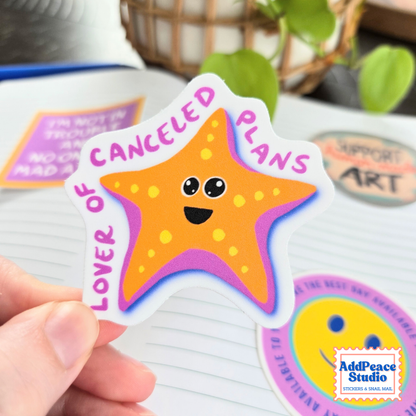A hand holds a die-cut sticker shaped like a smiling orange starfish with the text 'LOVER OF CANCELED PLANS' in pink. The sticker has a white border and is shown over a lined notebook with other colorful stickers in the background.