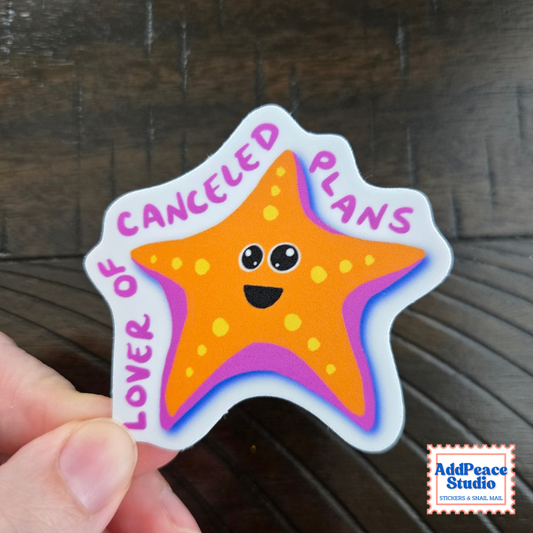 A hand holds a die-cut sticker featuring a smiling orange starfish with yellow spots, with the text 'LOVER OF CANCELED PLANS'. The sticker is photographed over a dark wood surface, with an 'AddPeace Studio' logo in the bottom right corner.