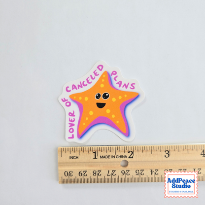 A die-cut sticker featuring a smiling orange starfish with the text 'LOVER OF CANCELED PLANS' in pink. The sticker is displayed above a wooden ruler on a plain white background, with an 'AddPeace Studio' watermark in the bottom right corner.